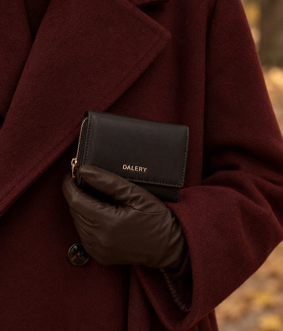 Main gantée de cuir tenant un portefeuille Dalery noir contre un manteau en laine bordeaux
