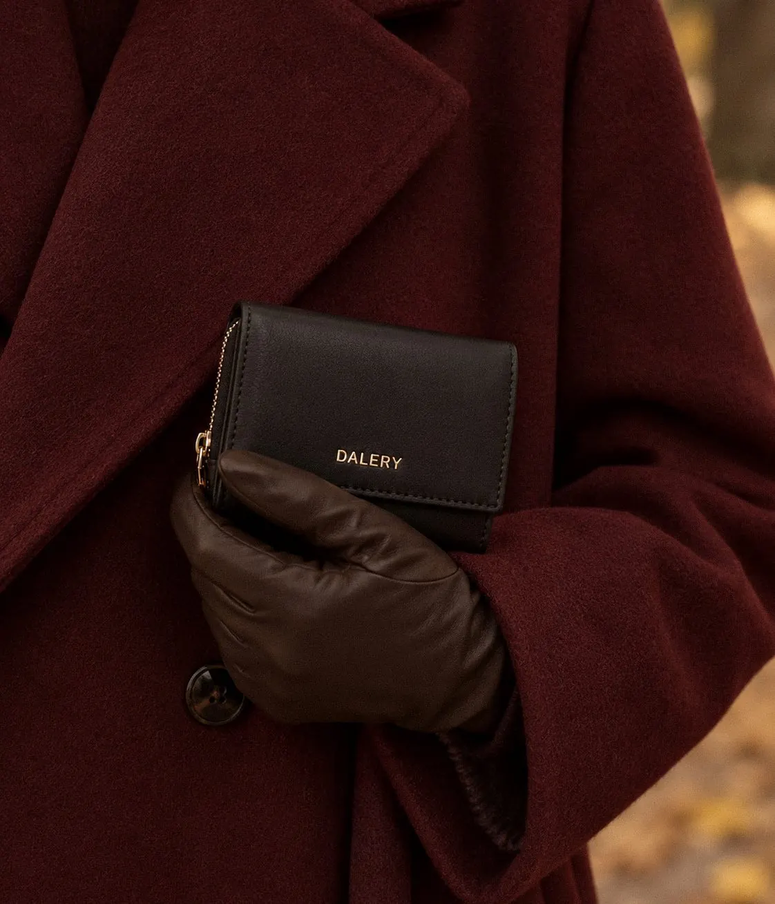 Main gantée de cuir tenant un portefeuille Dalery noir contre un manteau en laine bordeaux